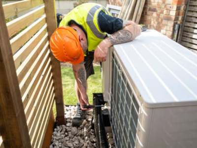 HVAC technician in an orange hard hat and high-visibility vest, installing or servicing an outdoor air conditioning unit.