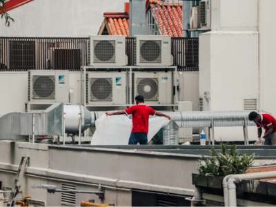 Two HVAC technicians in red shirts working on a rooftop commercial air conditioning equipment with multiple outdoor units visible in the background.