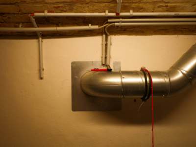 Galvanized metal HVAC duct with elbow connection mounted on a wall, with white PVC pipes running along the ceiling above.