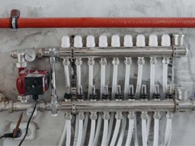 Chrome radiant floor heating manifold with multiple white PEX pipe connections, flow meters, and control valves mounted on the wall.
