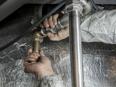 Overhead view of plumber's hands connecting brass and stainless steel pipe fittings on reflective insulated ductwork.