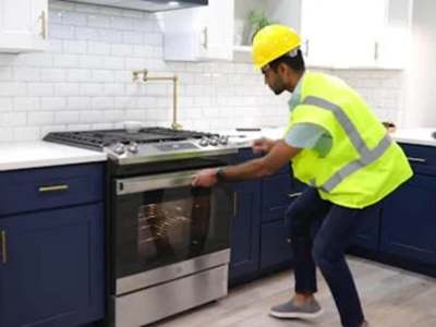 Worker in a yellow hard hat and safety vest installing a stainless steel stove in a modern kitchen.