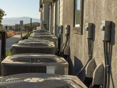 Row of outdoor HVAC condenser units with electrical disconnects along a stucco building wall, mountains in the background.