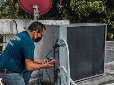 Technician in teal uniform and face mask using diagnostic gauges with blue hoses to service an outdoor unit on a rooftop.