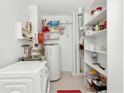 White utility room with washing machine, sink, water heater, wall shelves, and storage shelves holding cleaning supplies and tools.