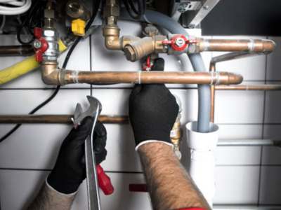 Plumber's hands in black gloves working on copper pipes and shut-off valves beneath a water heater or HVAC system.